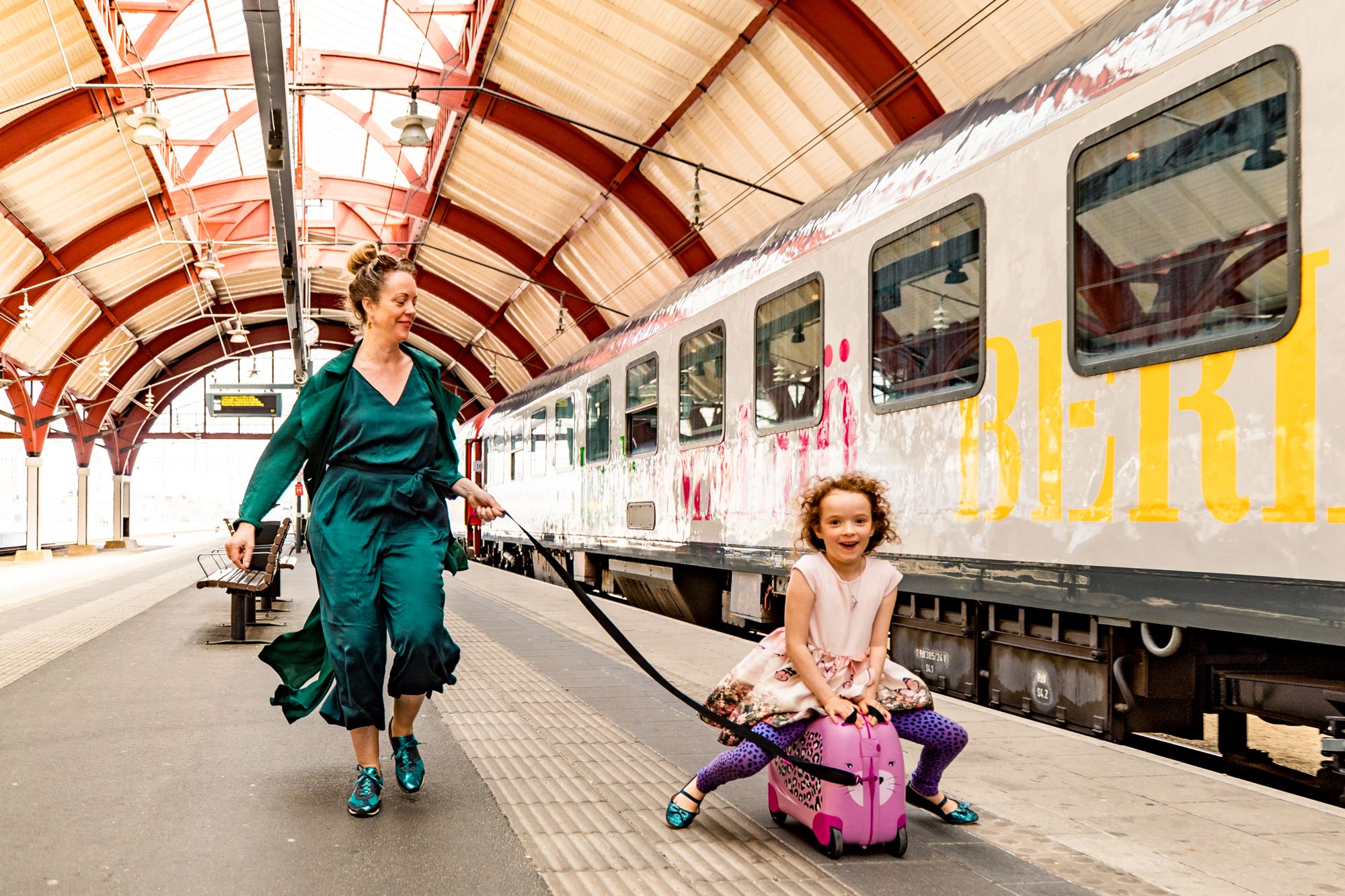 En kvinna drar ett glatt barn som sitter på en rosa resväska på en tågperrong.