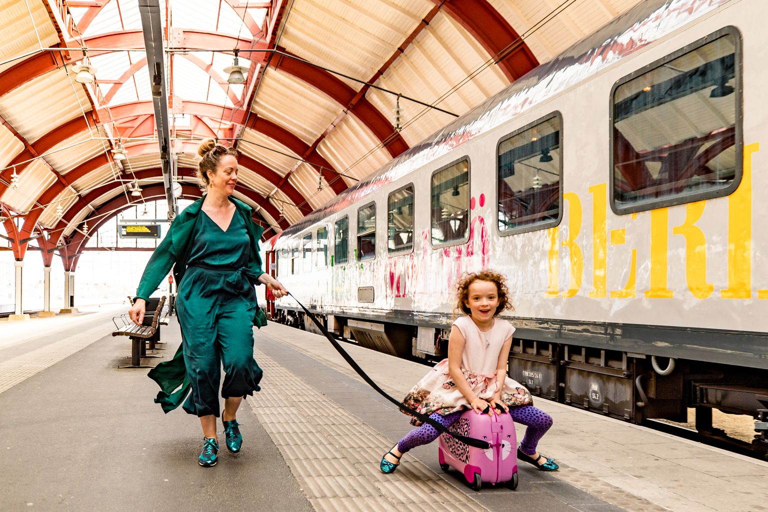 En kvinna drar ett glatt barn som sitter på en rosa resväska på en tågperrong.