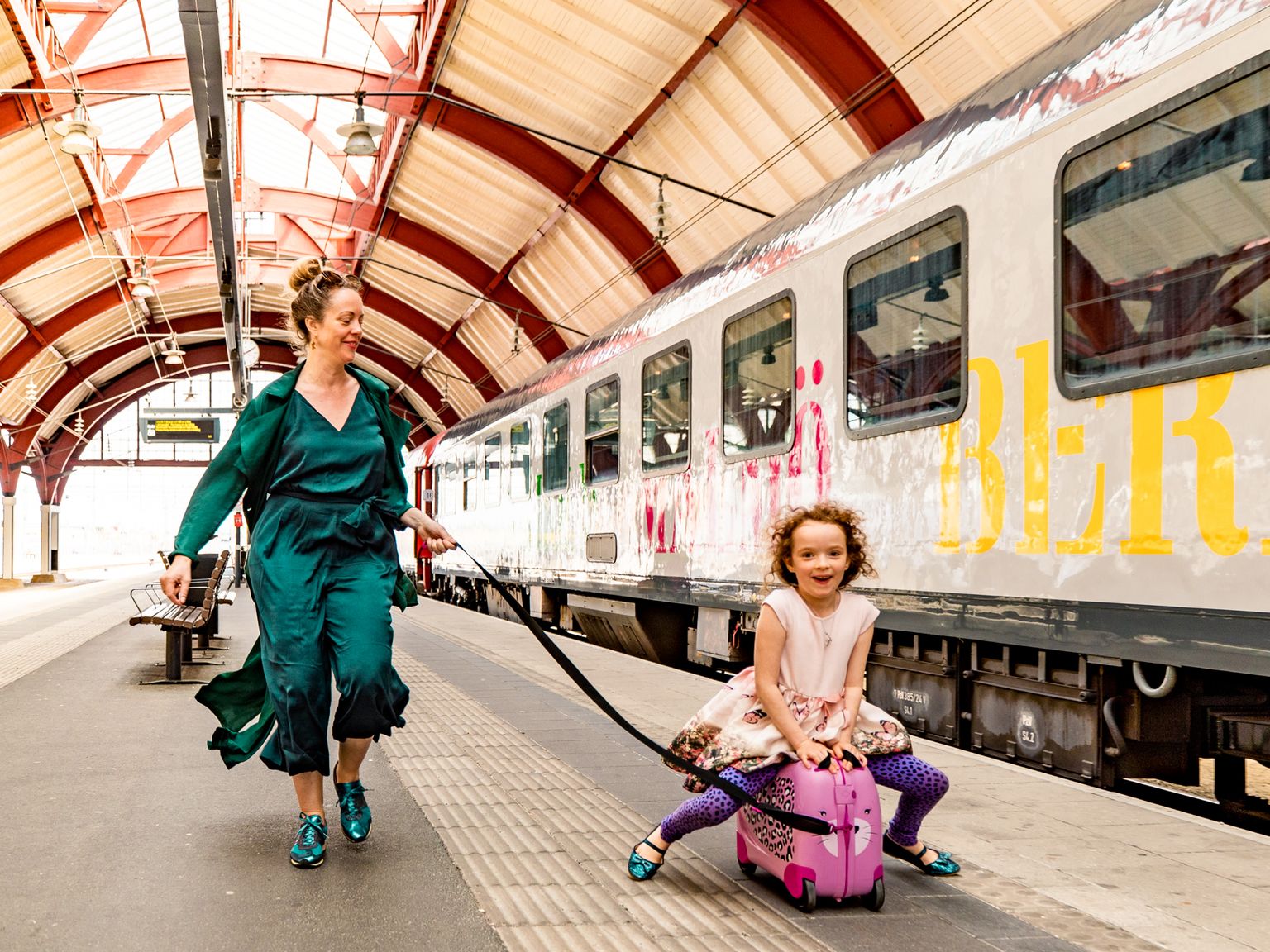 En kvinna drar ett glatt barn som sitter på en rosa resväska på en tågperrong.