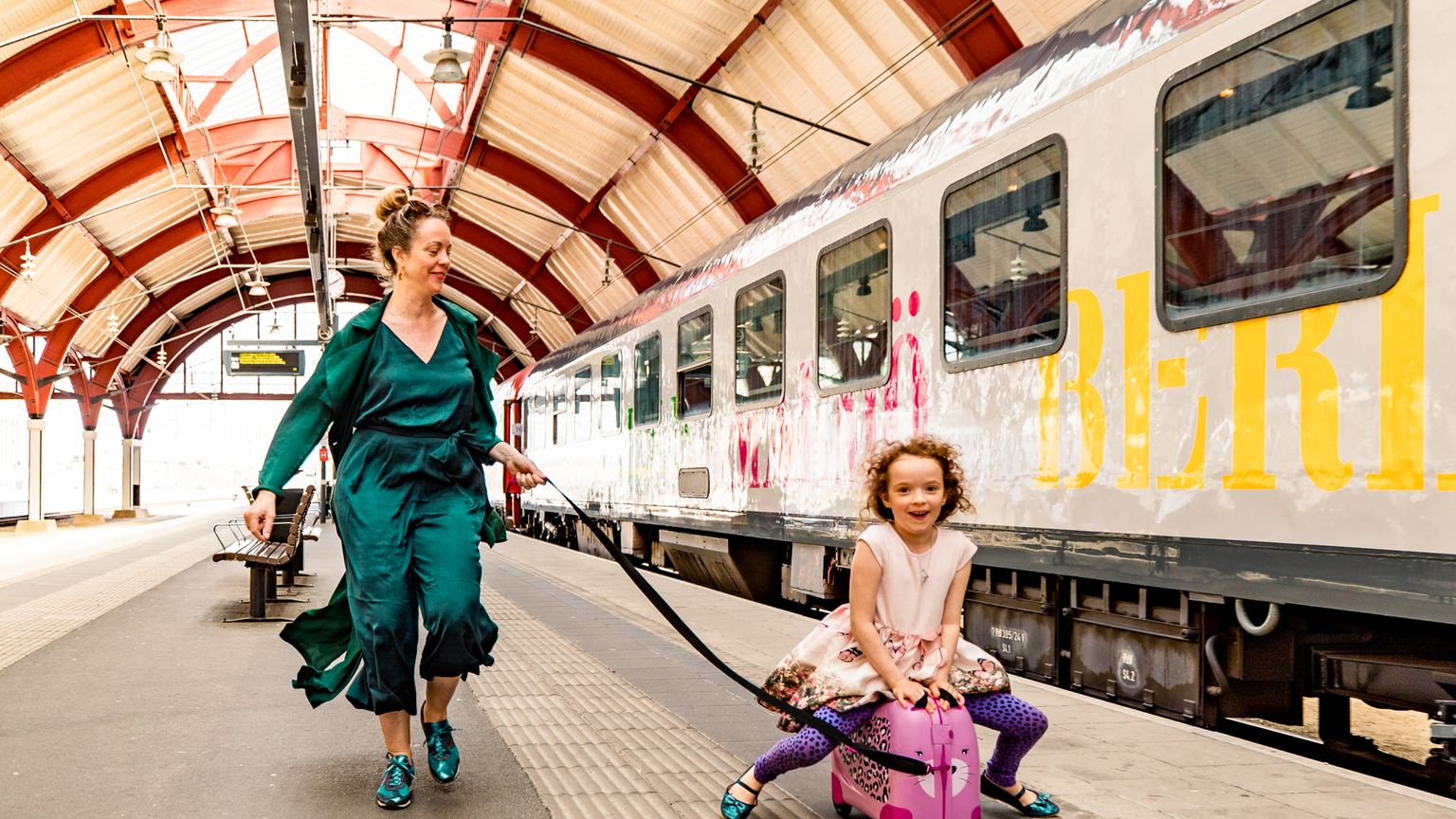 En kvinna drar ett glatt barn som sitter på en rosa resväska på en tågperrong.