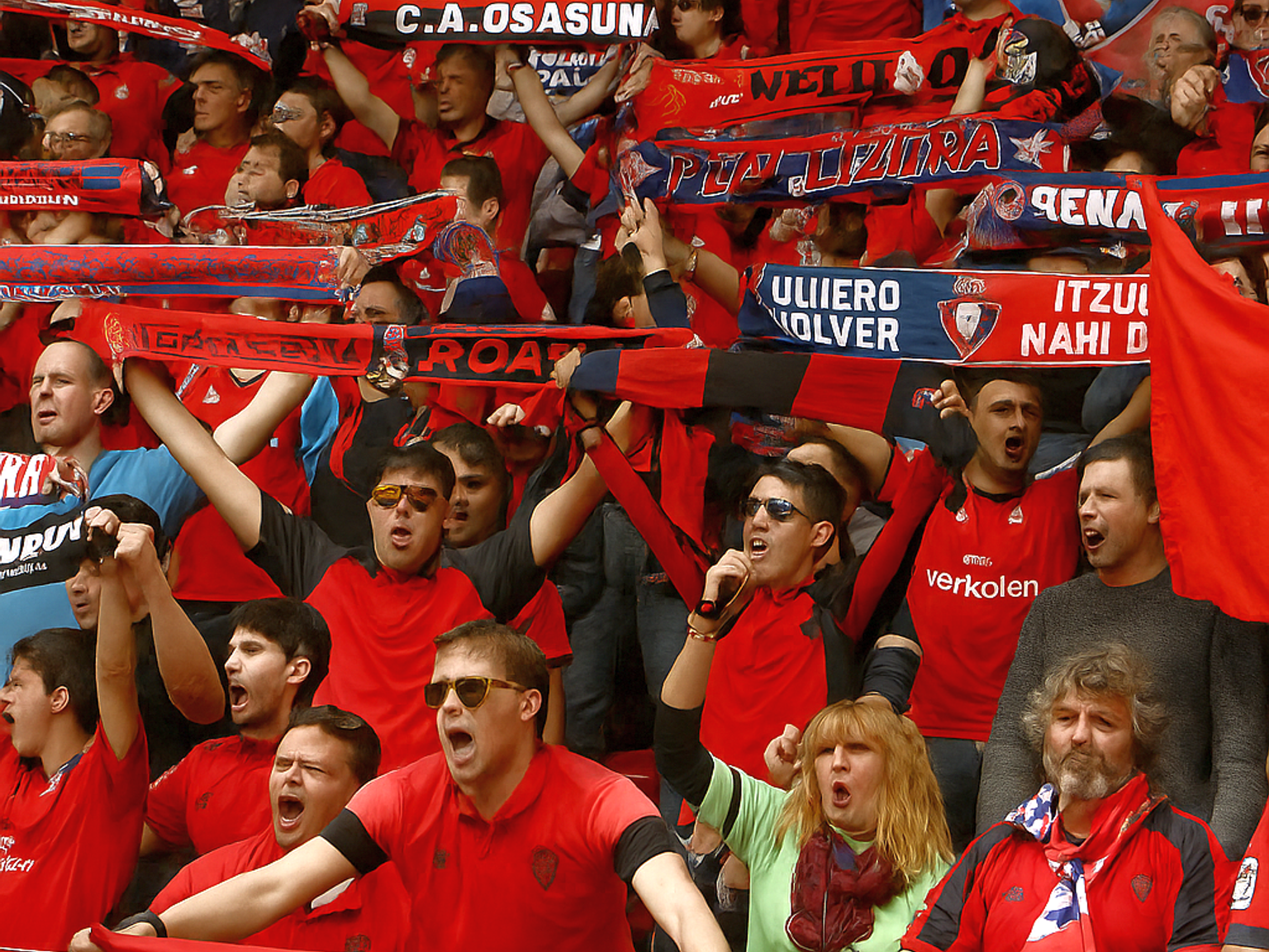 Enthousiaste voetbalfans in rood die juichen en met sjaals en vlaggen zwaaien, sommigen met "C.A. Osasuna" en het wapen van Navarra.