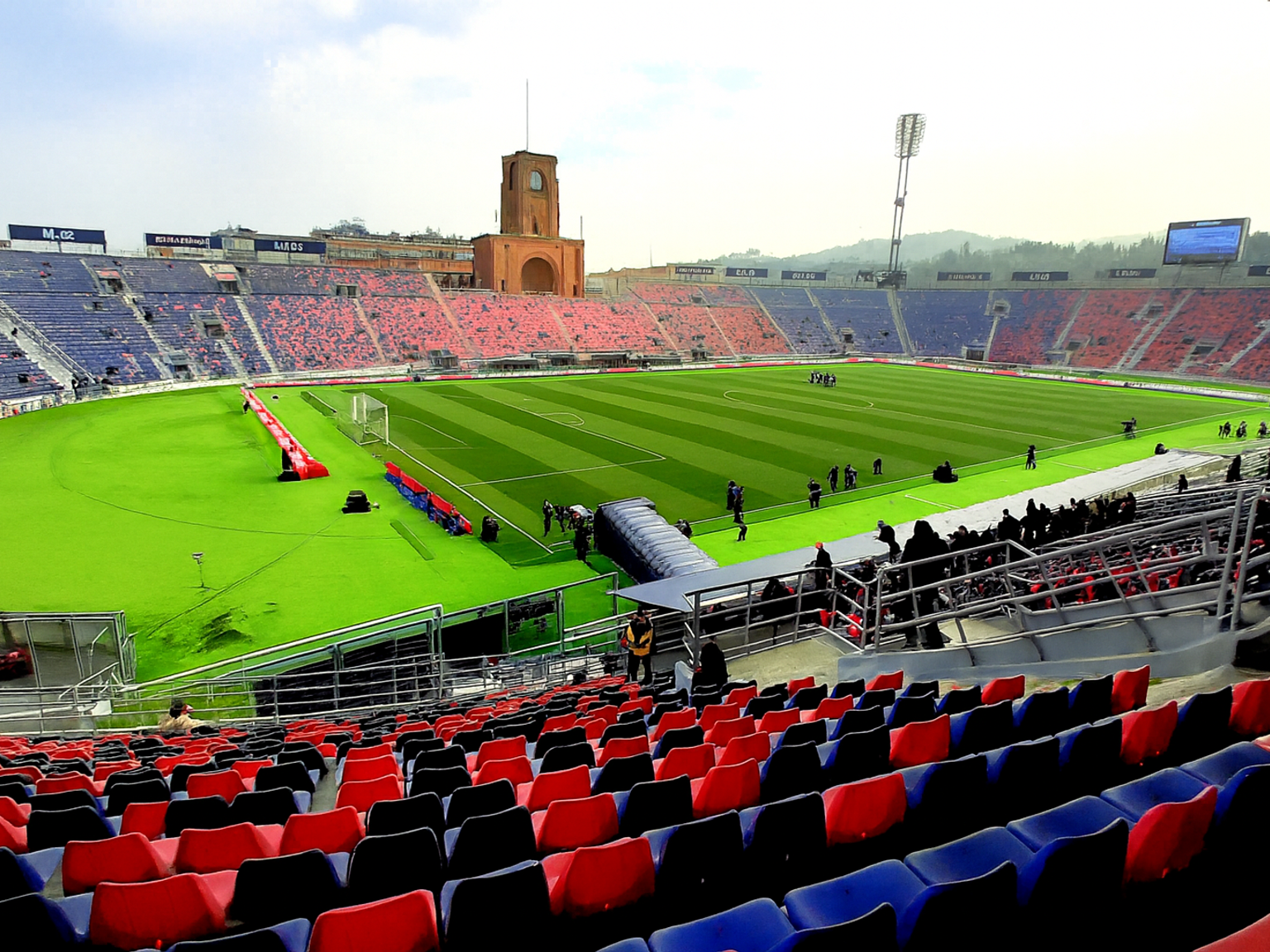 Un stade de football en grande partie vide avec un terrain vert, des sièges rouges et bleus, et une tour d'horloge distinctive.
