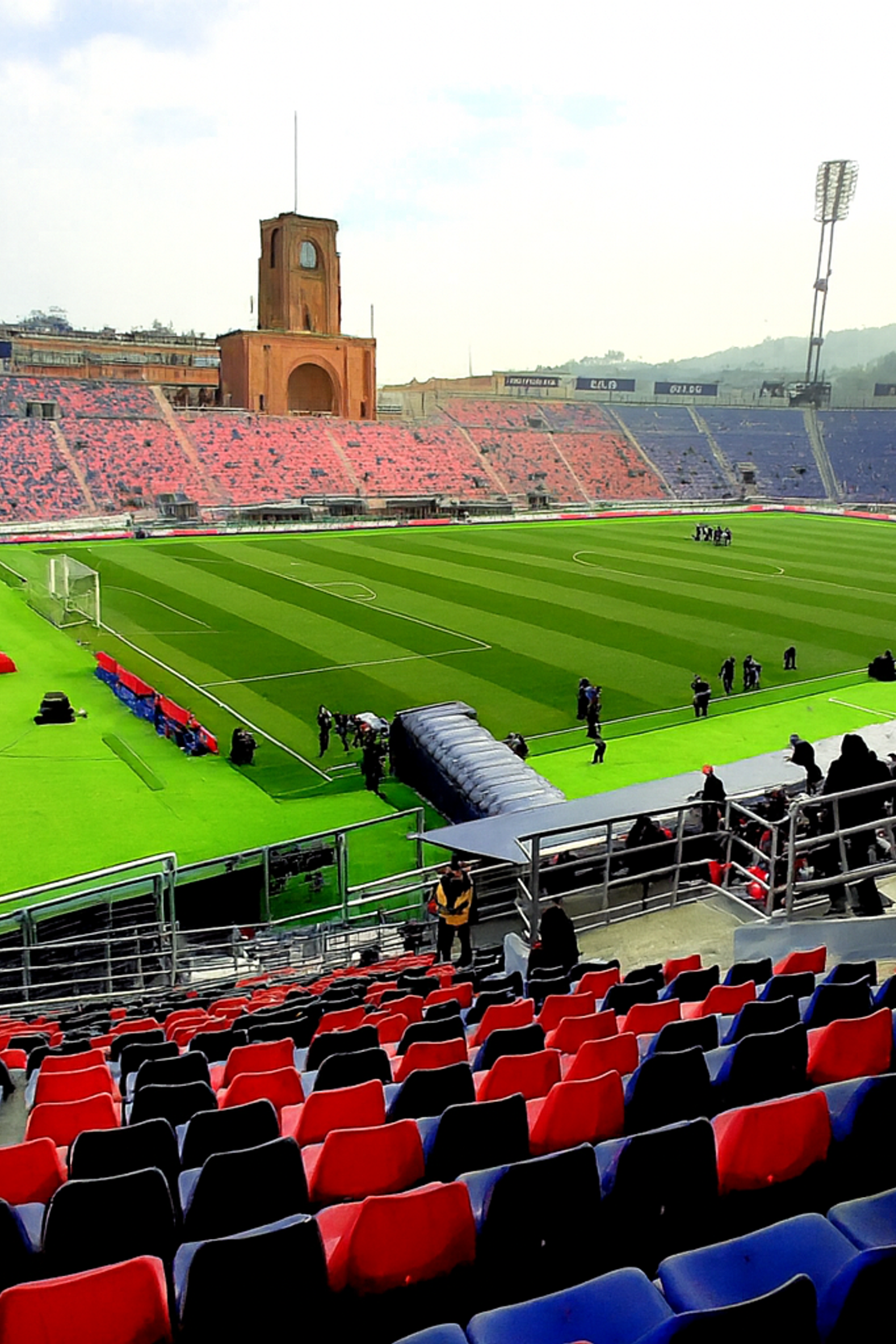 Un stade de football en grande partie vide avec un terrain vert, des sièges rouges et bleus, et une tour d'horloge distinctive.