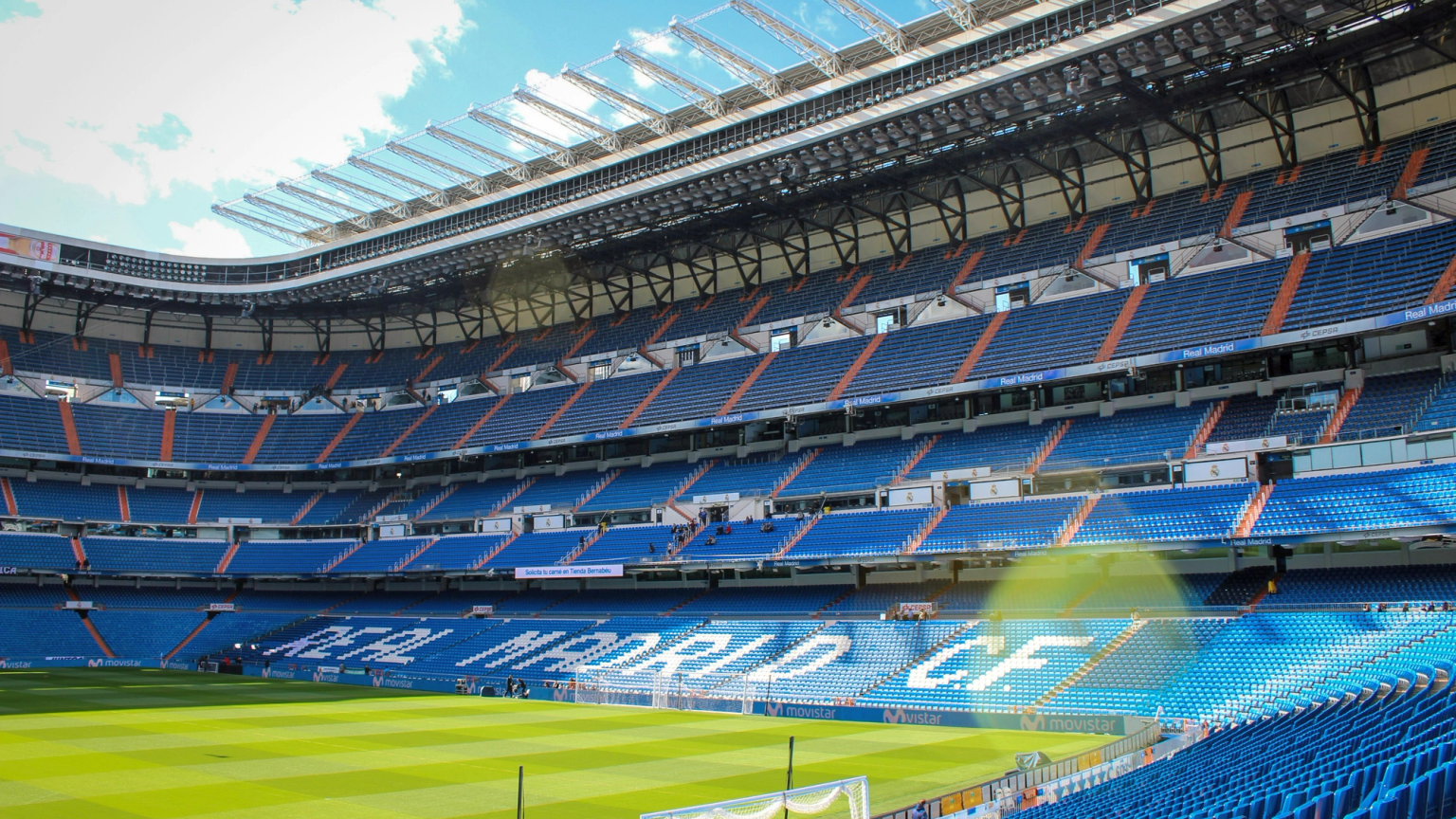 Leeres Real Madrid C.F. Fußballstadion mit blauen Sitzen und einem grünen Spielfeld unter blauem Himmel.