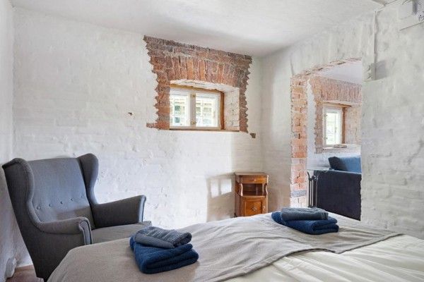 Et rustikt soveværelse med hvide vægge, blotlagt murværk omkring vinduerne, en seng med blå håndklæder og en grå lænestol.