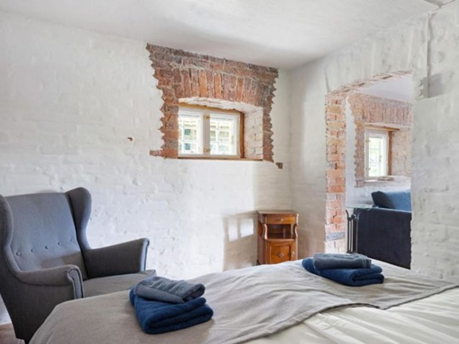 Et rustikt soveværelse med hvide vægge, blotlagt murværk omkring vinduerne, en seng med blå håndklæder og en grå lænestol.
