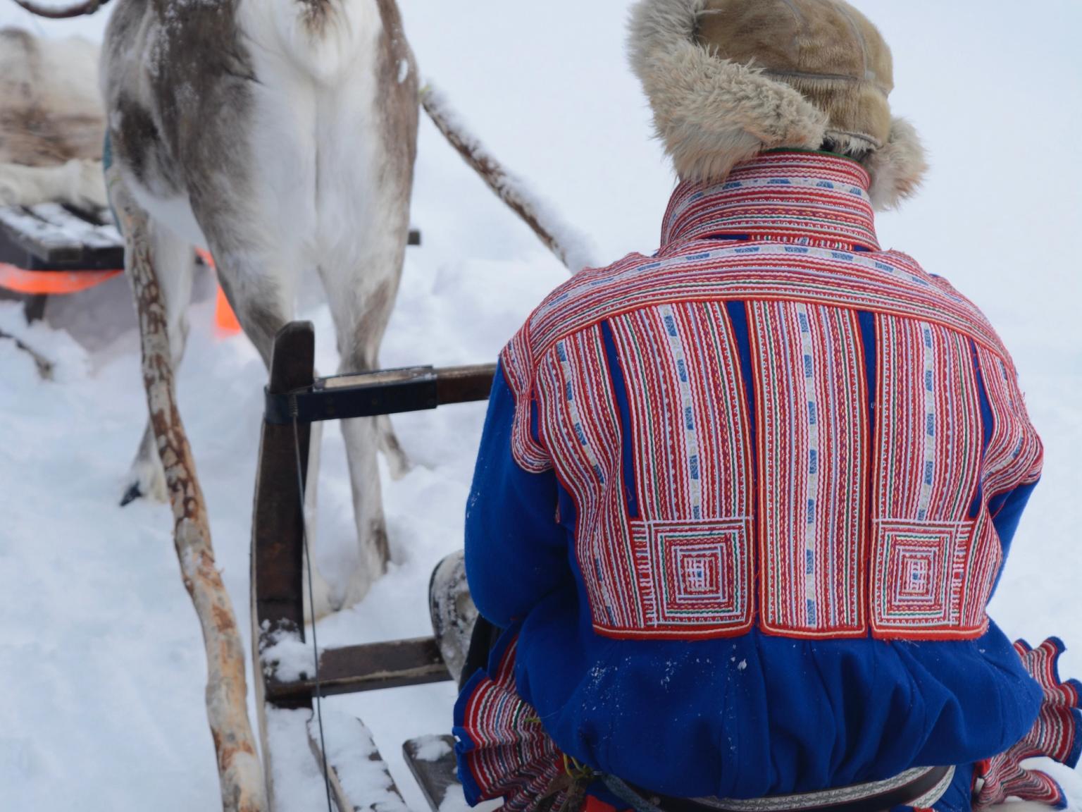 en mand i en blå og rød jakke sidder på en slæde trukket af rensdyr
