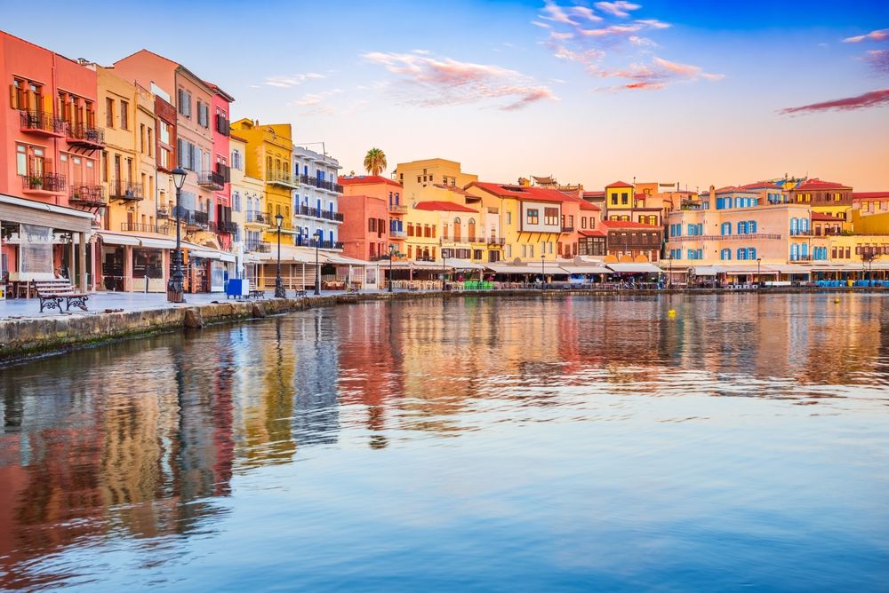 Farverige bygninger, der spejles i en rolig havn ved solnedgang.