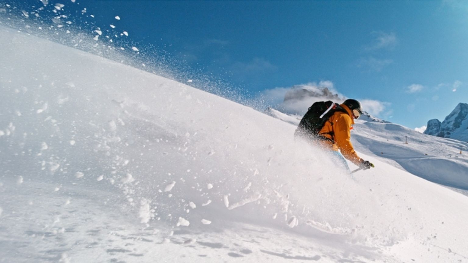 Skier in an orange jacket kicking up snow on a sunny mountain slope.