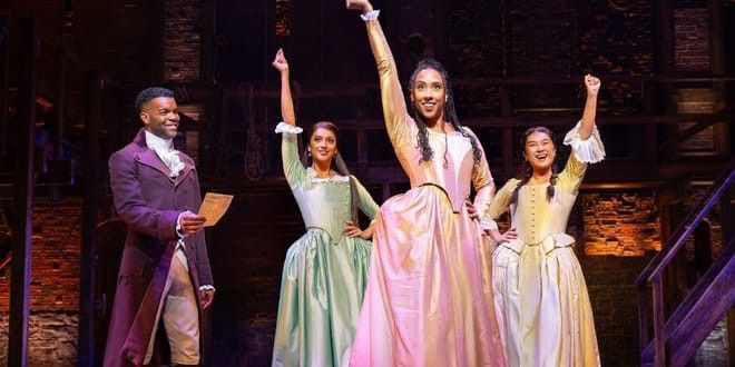 Fyra artister från "Hamilton" på scen i tidsenliga kostymer, en man och tre kvinnor, där två av kvinnorna lyfter armarna. Musikaler - Hamilton - London