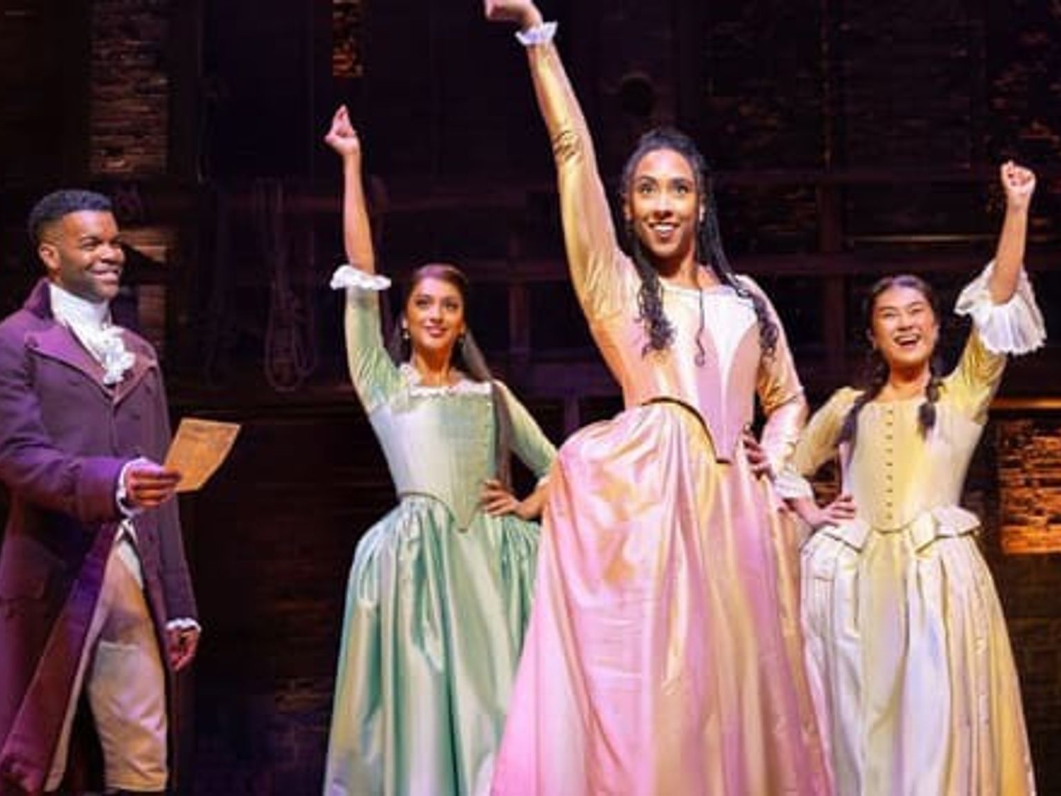 Fyra artister från "Hamilton" på scen i tidsenliga kostymer, en man och tre kvinnor, där två av kvinnorna lyfter armarna. Musikaler - Hamilton - London
