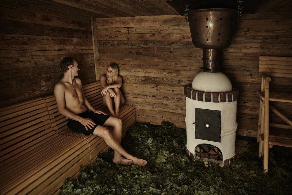 En man och en kvinna kopplar av på en träbänk i en rustik bastu med en kamin, en koppargryta och löv som täcker golvet.