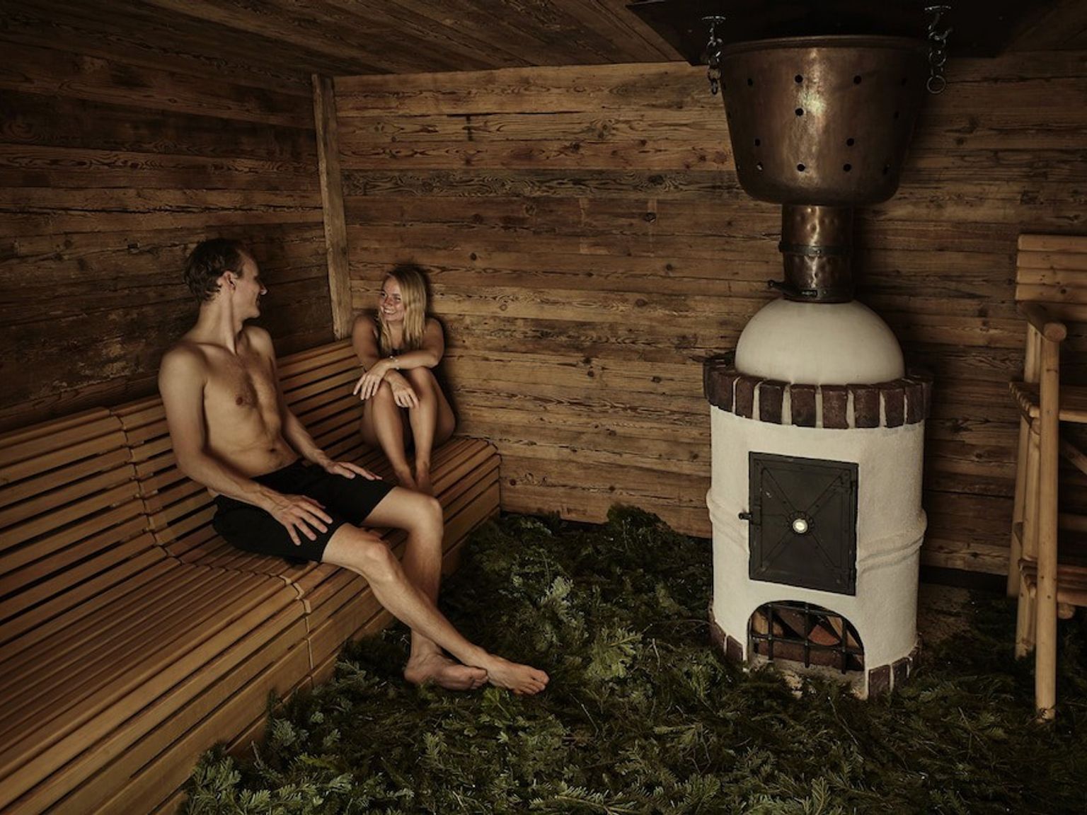 En man och en kvinna kopplar av på en träbänk i en rustik bastu med en kamin, en koppargryta och löv som täcker golvet.