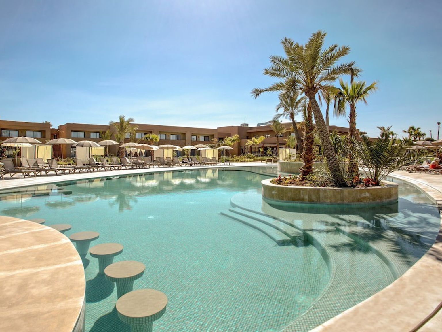 Et moderne feriested svømmebasseng med en swim-up bar, en palmeøy, solsenger og en hotellbygning under en klar blå himmel.