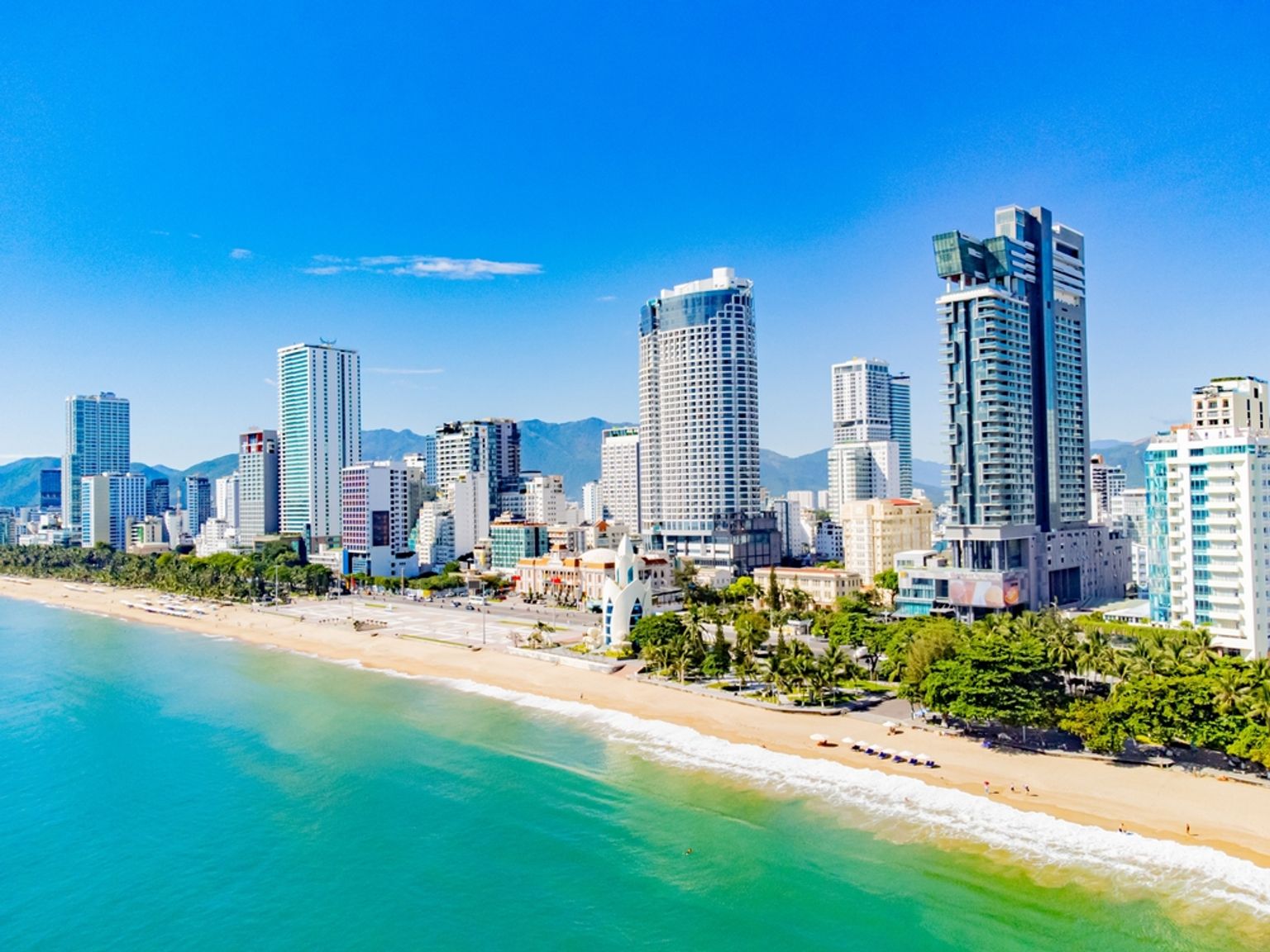 Flygfoto av en livlig stadsstrand med turkost vatten, sandstrand, palmer och en skyline av moderna höghus. Nha Trang, Vietnam