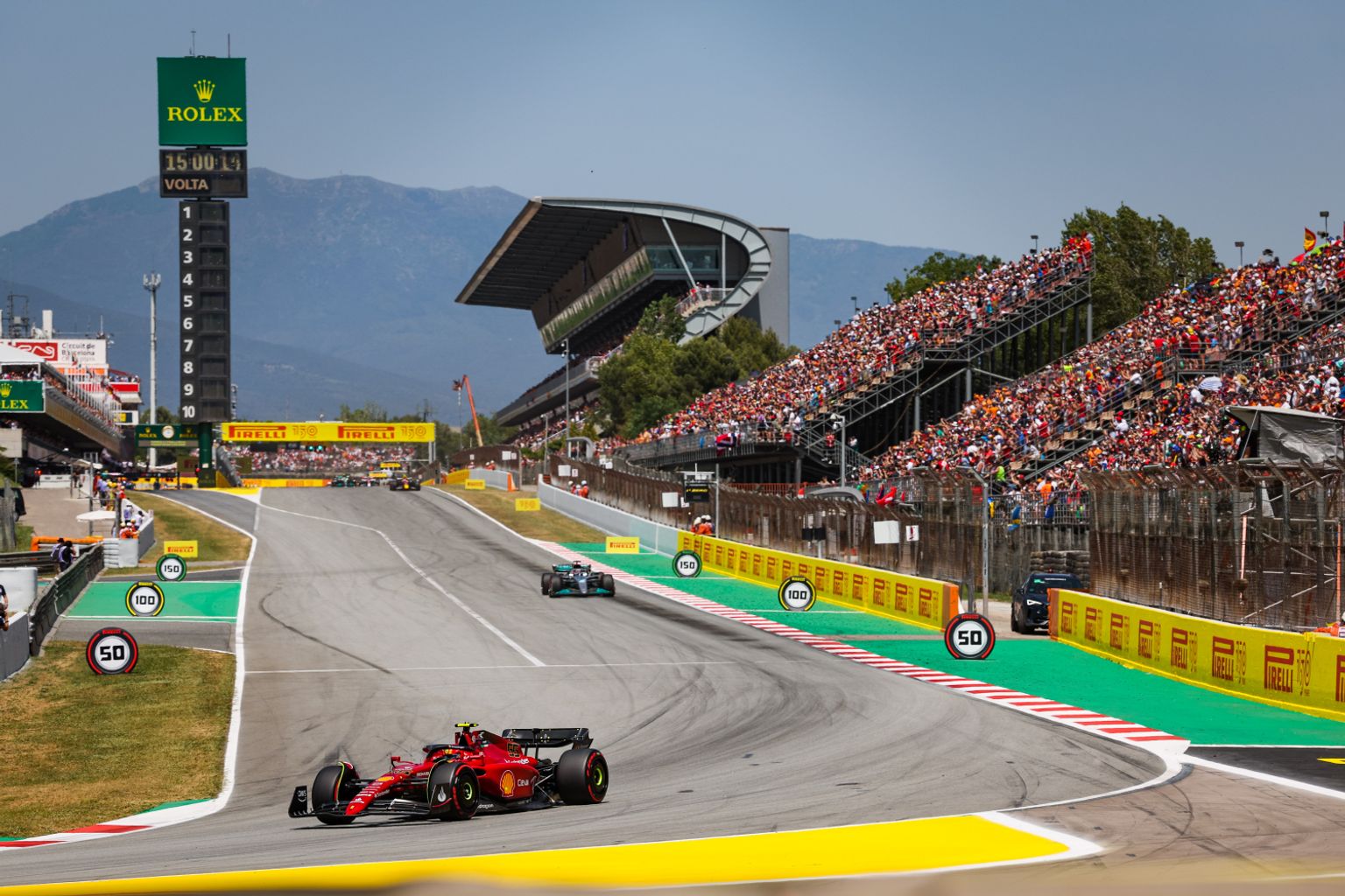 A red Formula 1 car races on a track with a green car, crowded grandstands, mountains, and a Rolex timing tower.