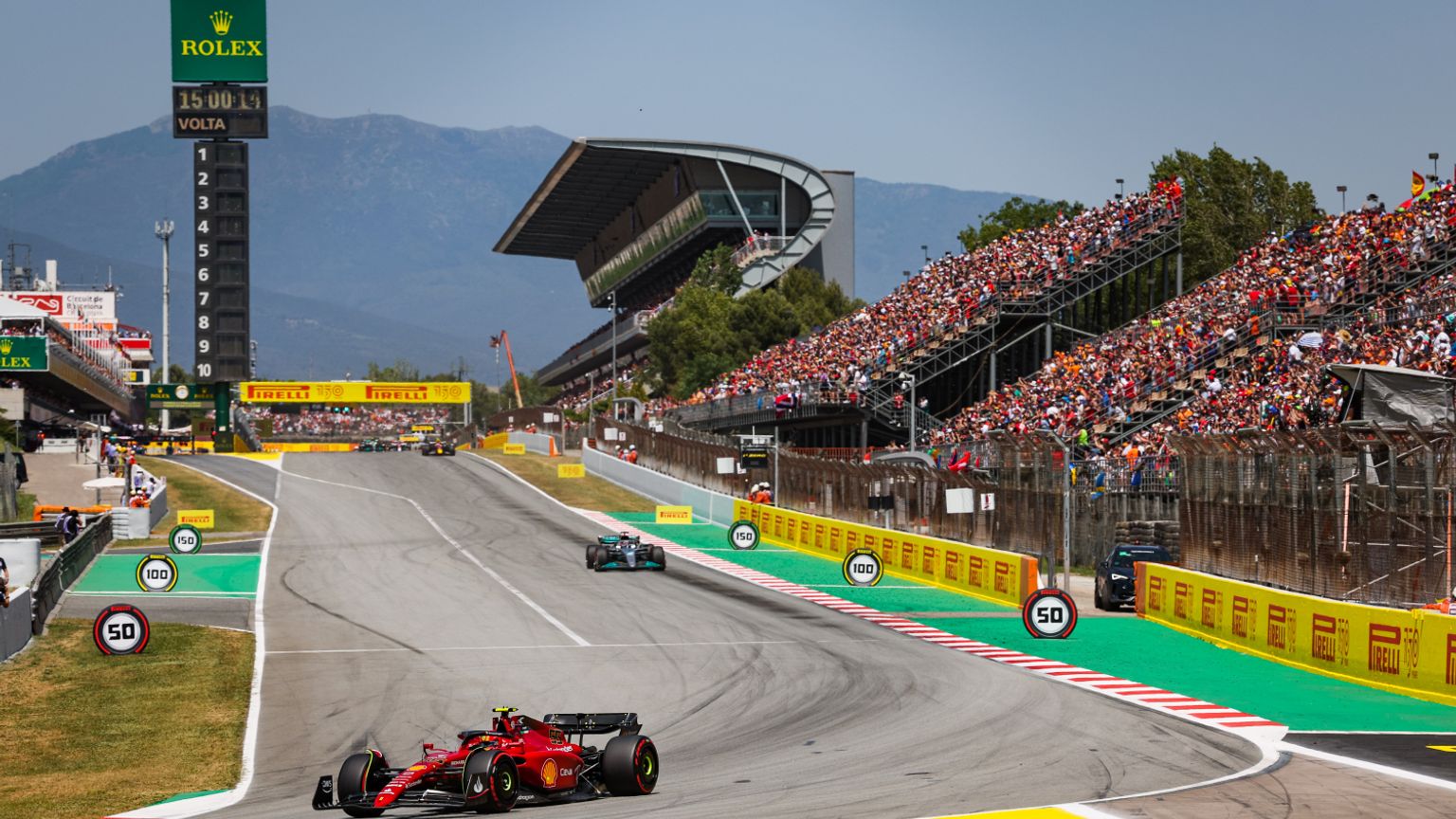 A red Formula 1 car races on a track with a green car, crowded grandstands, mountains, and a Rolex timing tower.