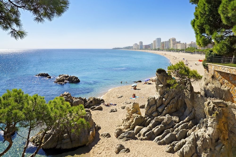 Solrig sandstrand med turkis vand, klippeområder og en bysilhuet. Playa de Aro, Spanien