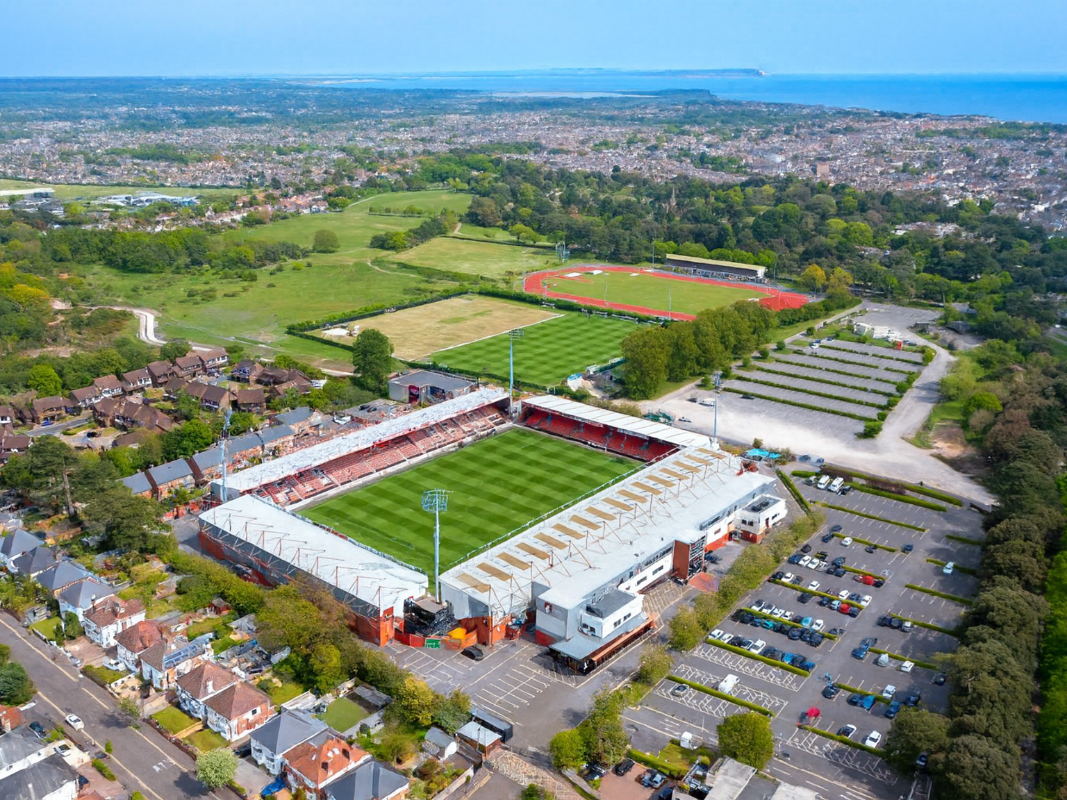Luftaufnahme eines Fußballstadions mit angrenzenden Sportplätzen, Parkplätzen und Wohngebäuden, mit Blick auf eine Küstenstadt.