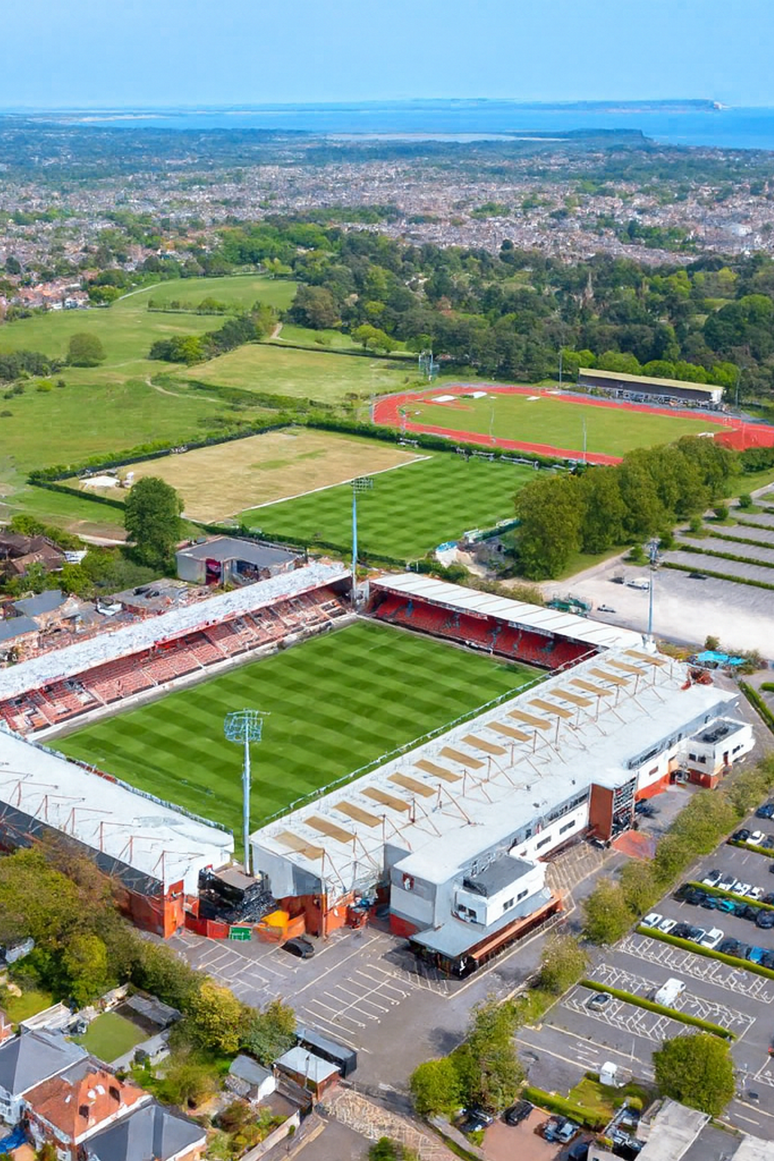 Luftaufnahme eines Fußballstadions mit angrenzenden Sportplätzen, Parkplätzen und Wohngebäuden, mit Blick auf eine Küstenstadt.