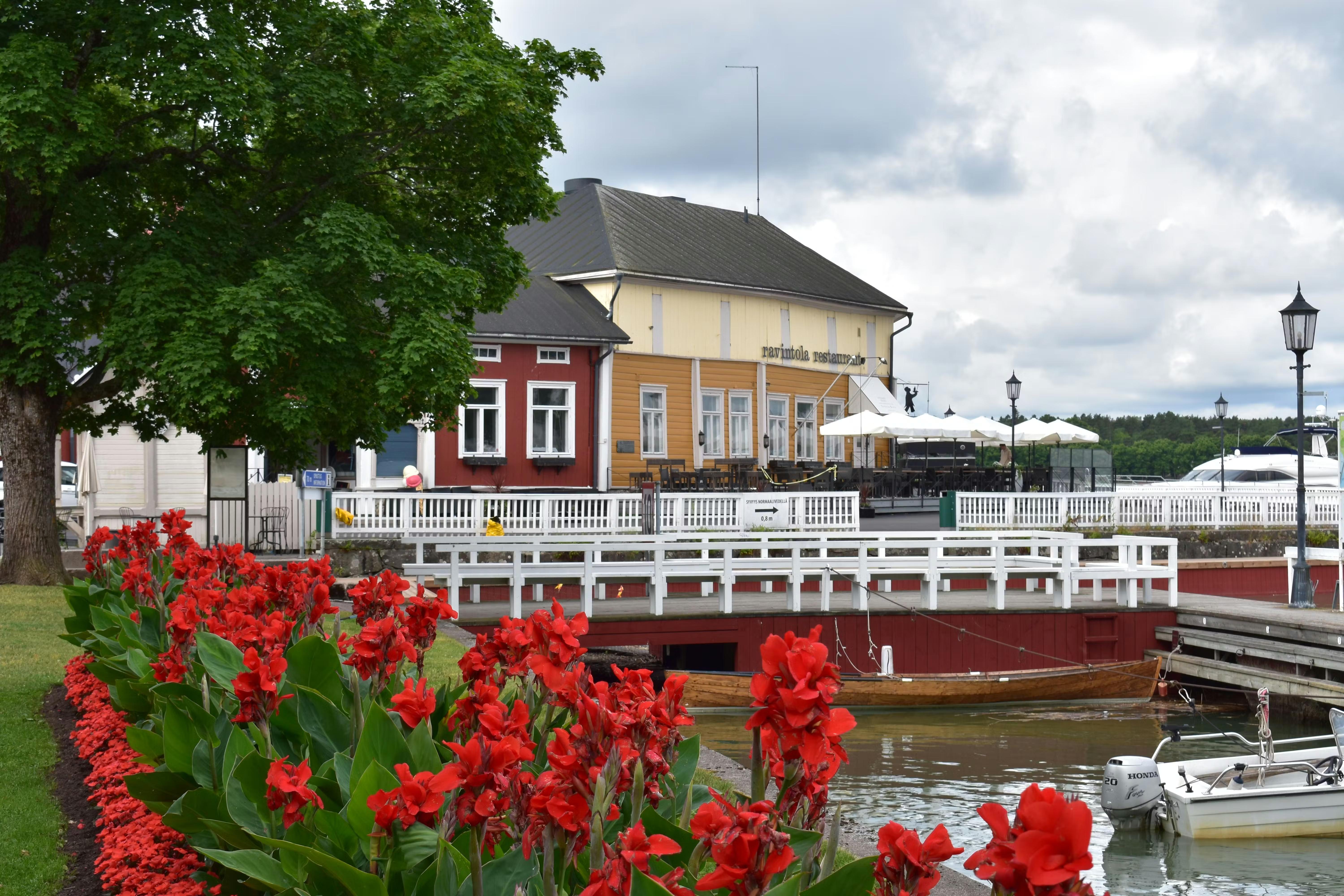 Røde blomster pryder en havnefront med farverige bygninger, både og en overskyet himmel.