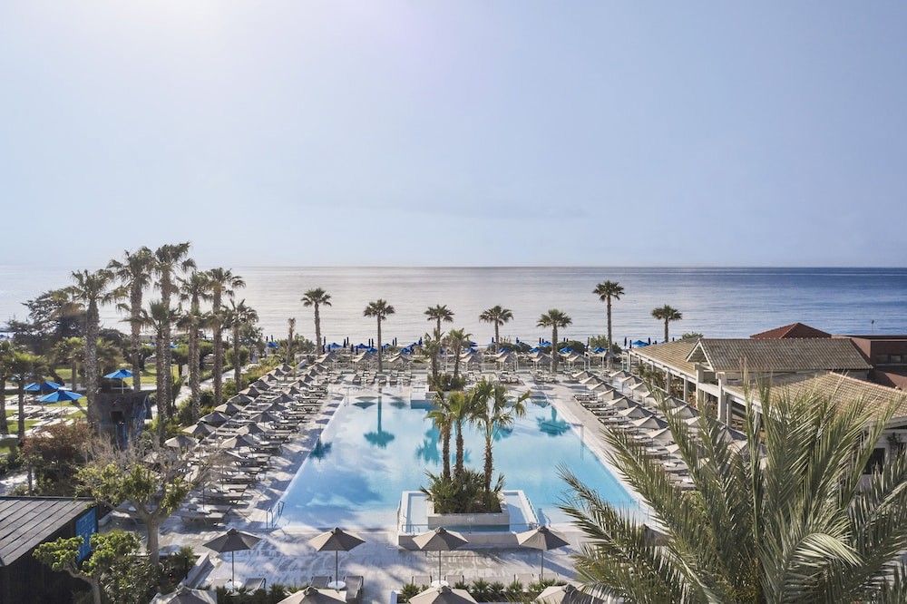 Resort pool with palm trees, lounge chairs, and ocean view under a clear sky. Esperides Beach Resort