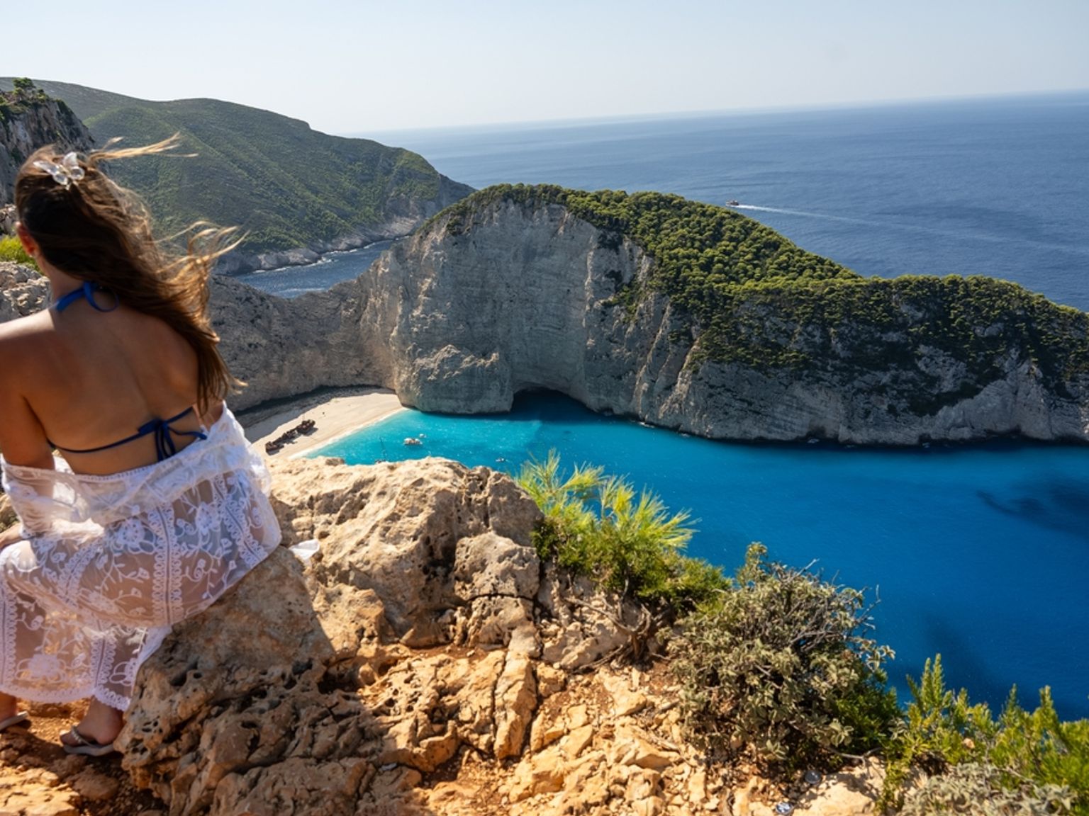 En kvinna i en vit spetskaftan sitter på en klippig klippa och blickar ut över en fantastisk turkos vik med en sandstrand inbäddad mellan höga vita klippor.