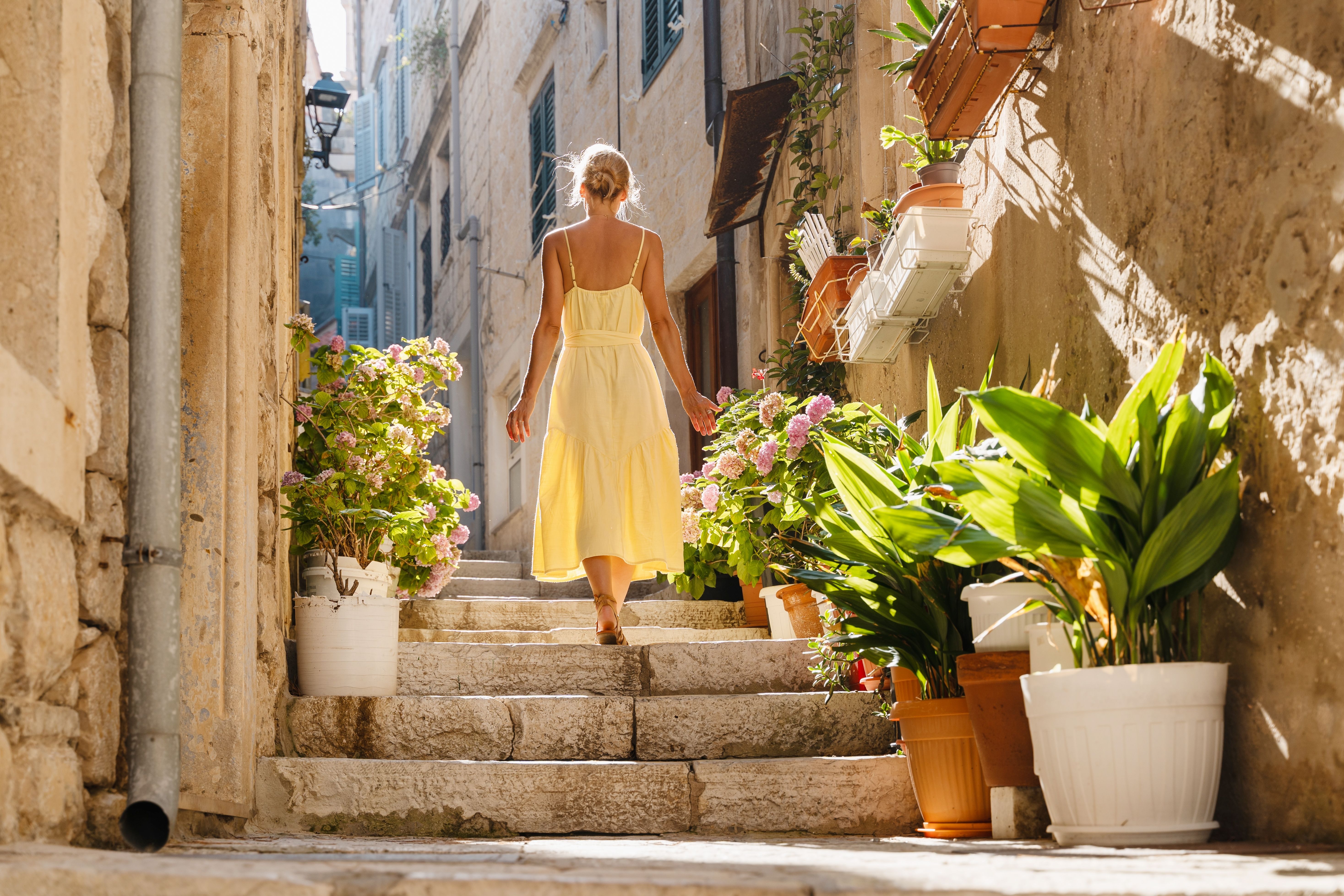 Vrouw in een gele jurk loopt een zonovergoten stenen trap op in een smalle steeg met potplanten. Korčula, Kroatië