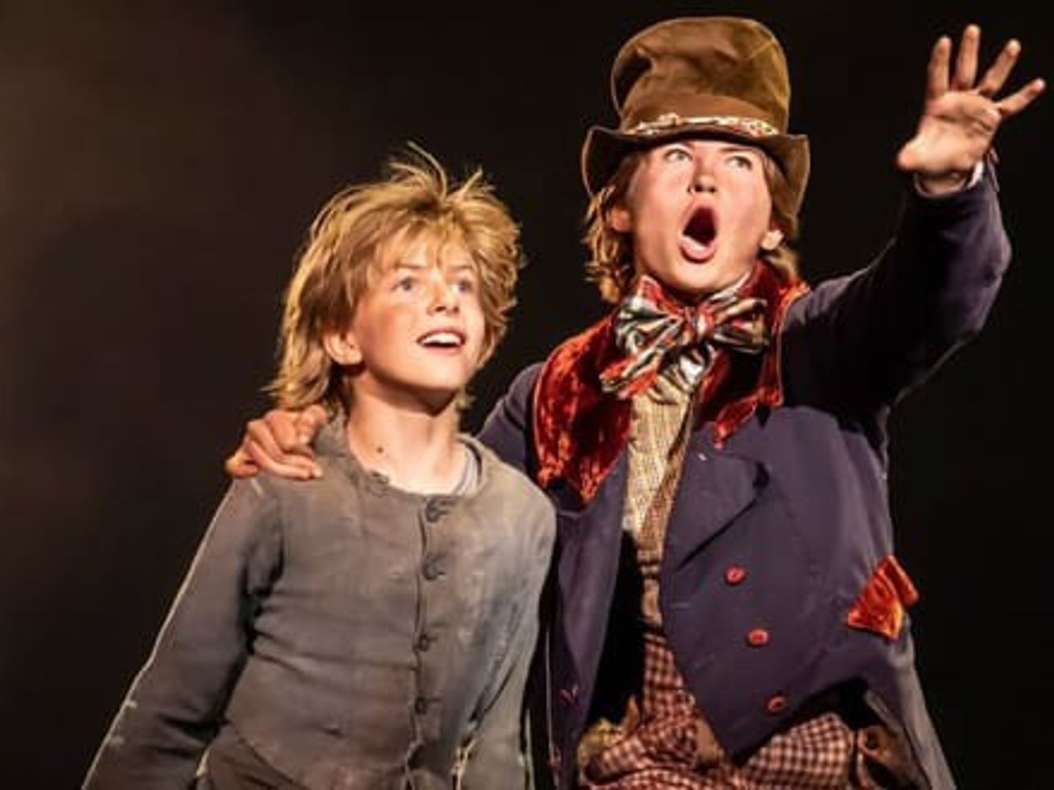 Two boys in theatrical costumes on a dark stage. One in ragged clothes looks up, while the other in a top hat and colorful suit sings with an arm raised.