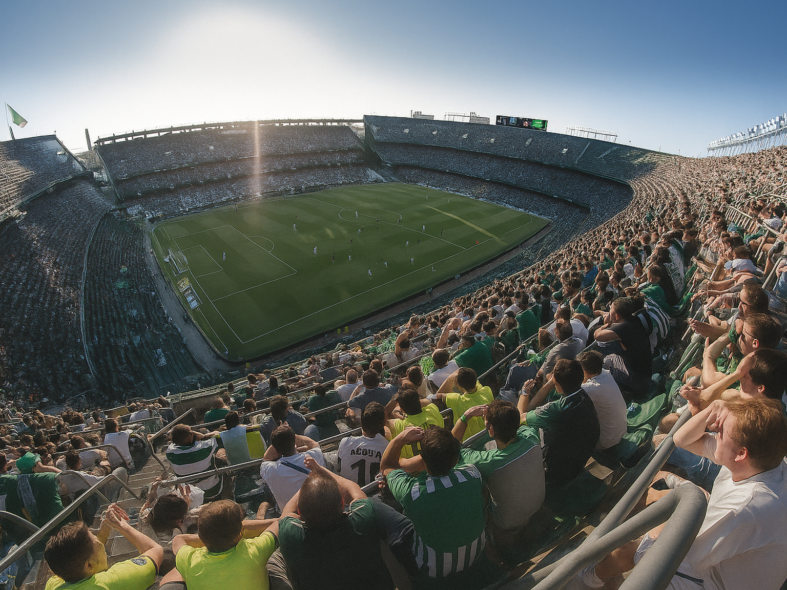 Groothoekzicht op een voetbalstadion vol fans in groen en wit onder fel zonlicht.