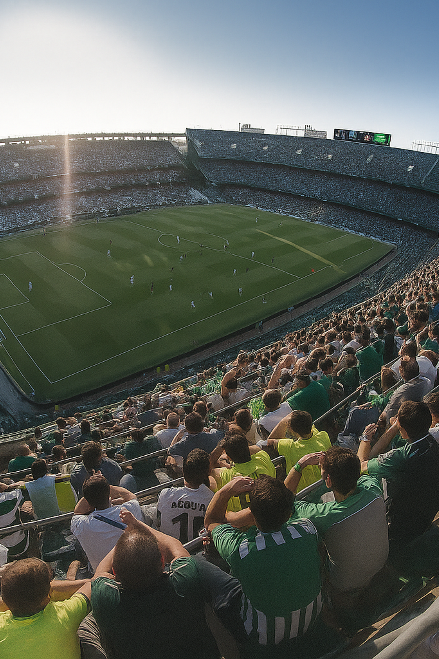 Groothoekzicht op een voetbalstadion vol fans in groen en wit onder fel zonlicht.