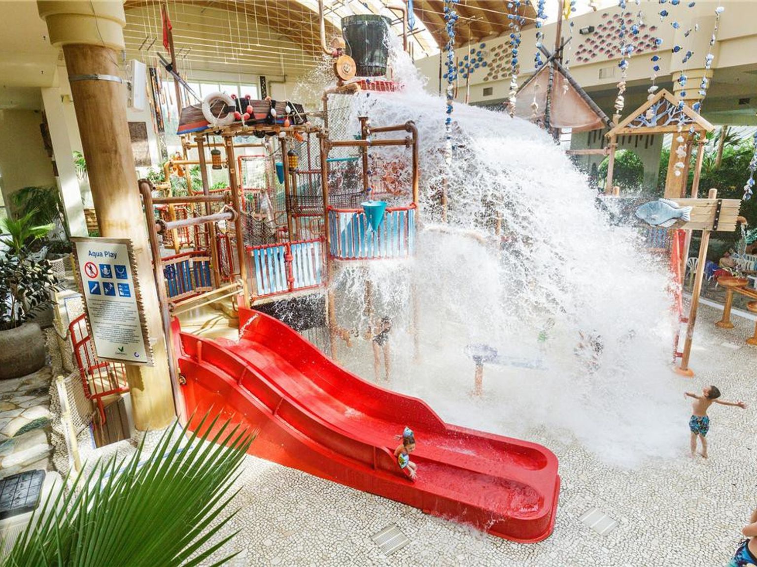 Overdekt waterpark met een grote emmer die water op een speelstructuur, rode glijbaan en kinderen stort. Center Parcs Park Hochsauerland