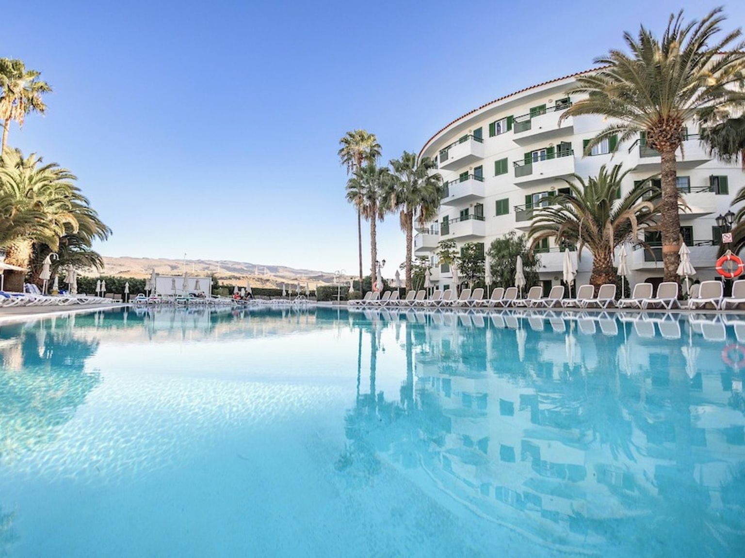 En klarblå swimmingpool speglar en vit hotellbyggnad och palmer under en solig himmel.