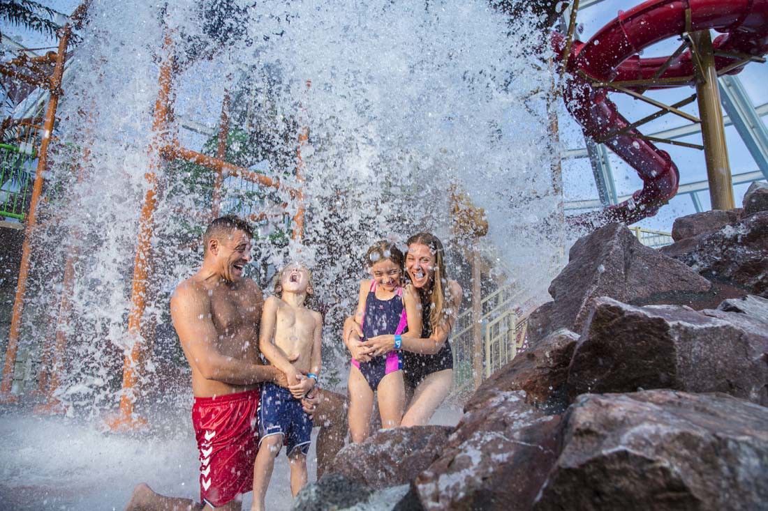 En familie på fire som ler under en stor vannstråle i en vannpark. Lalandia Rødby