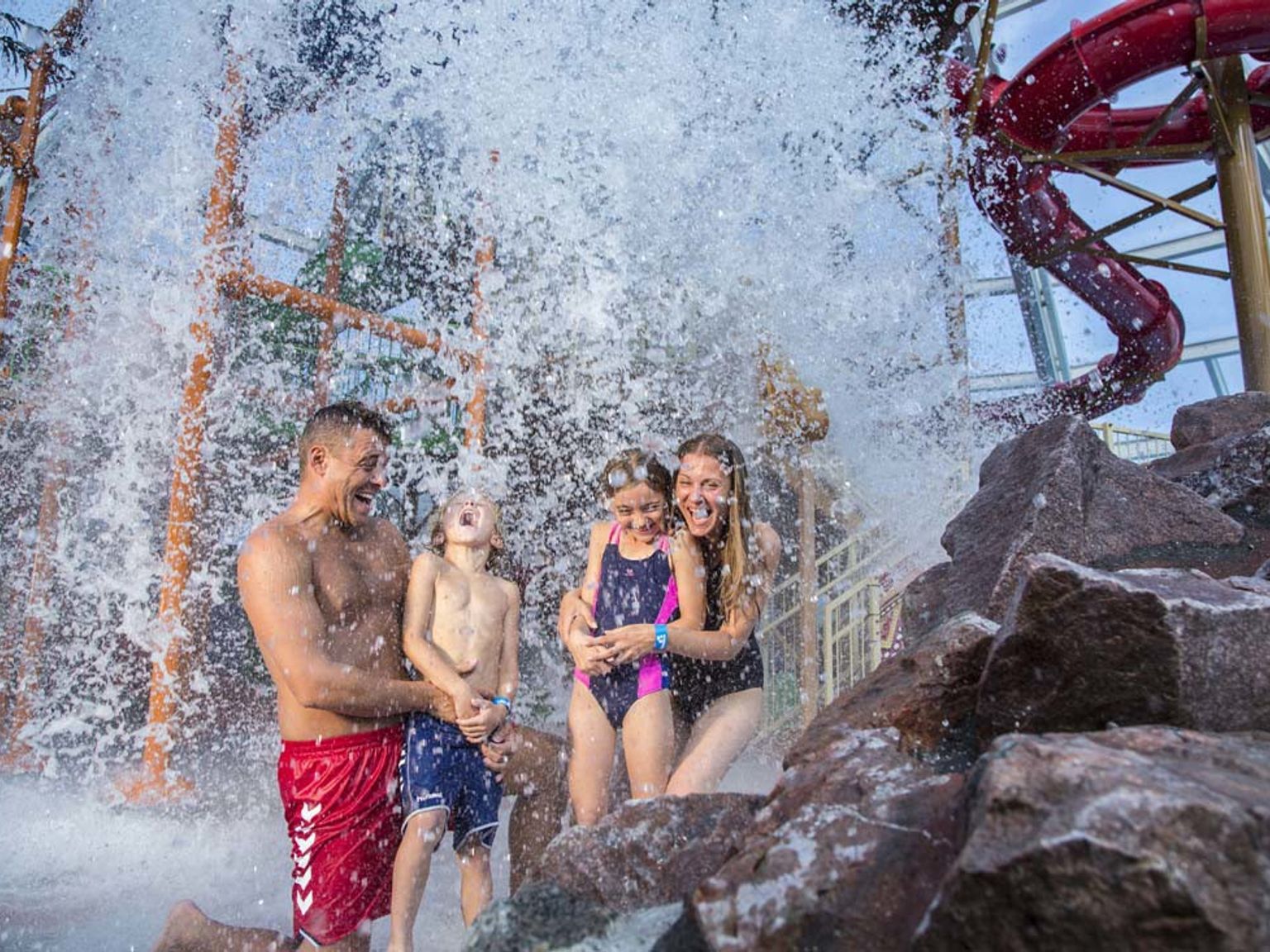En familie på fire som ler under en stor vannstråle i en vannpark. Lalandia Rødby