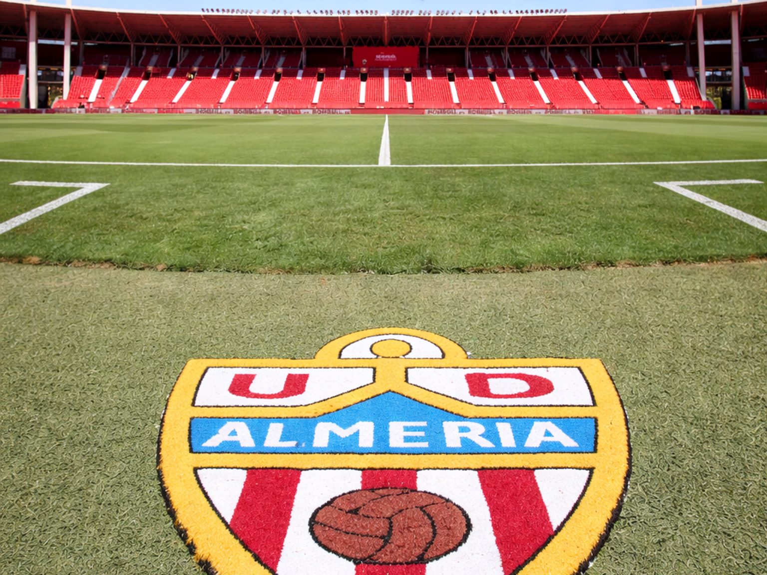 U.D.-Almería-Logo auf einem Fußballfeld, mit leeren roten Stadionsitzen unter einem klaren blauen Himmel.