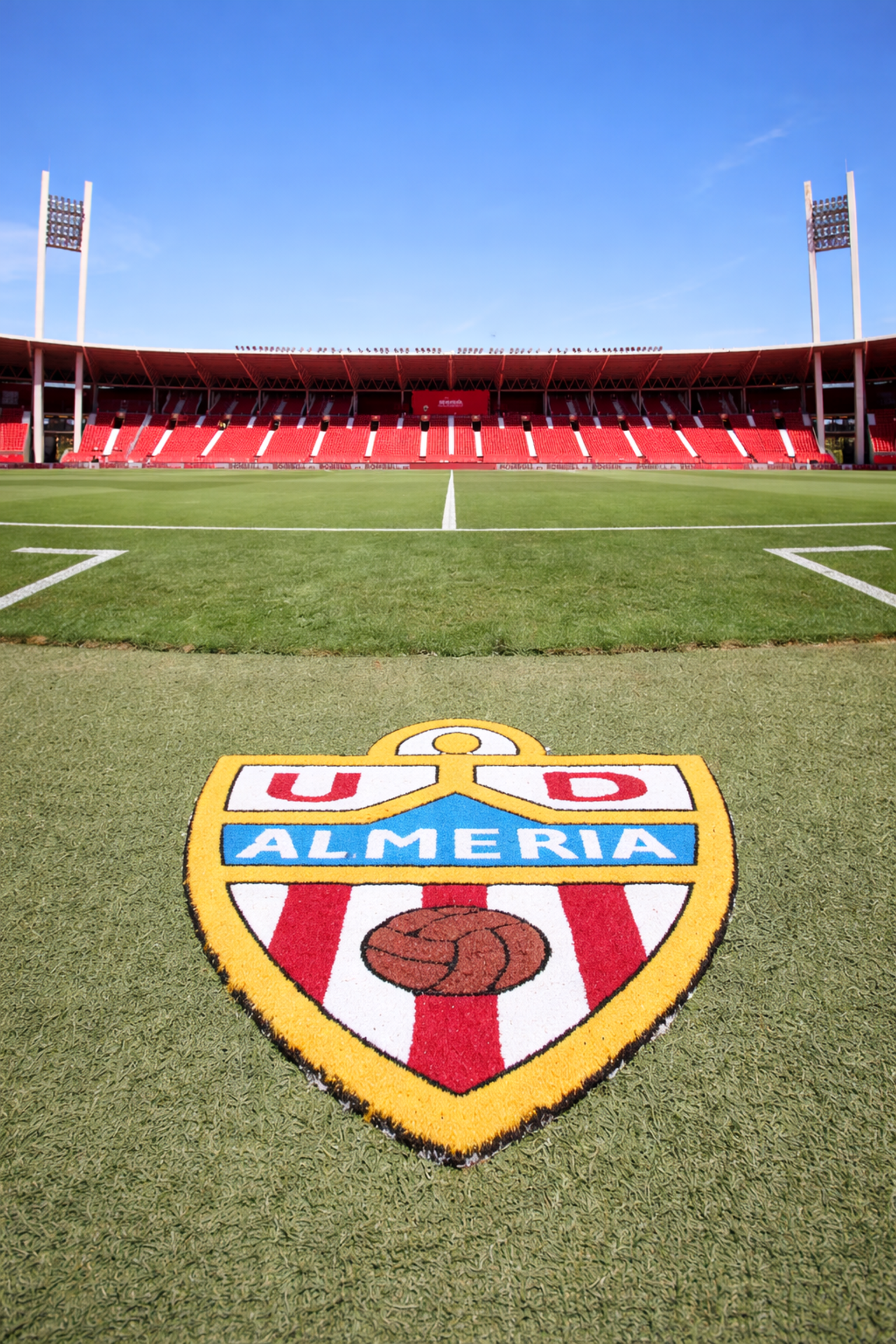 U.D.-Almería-Logo auf einem Fußballfeld, mit leeren roten Stadionsitzen unter einem klaren blauen Himmel.