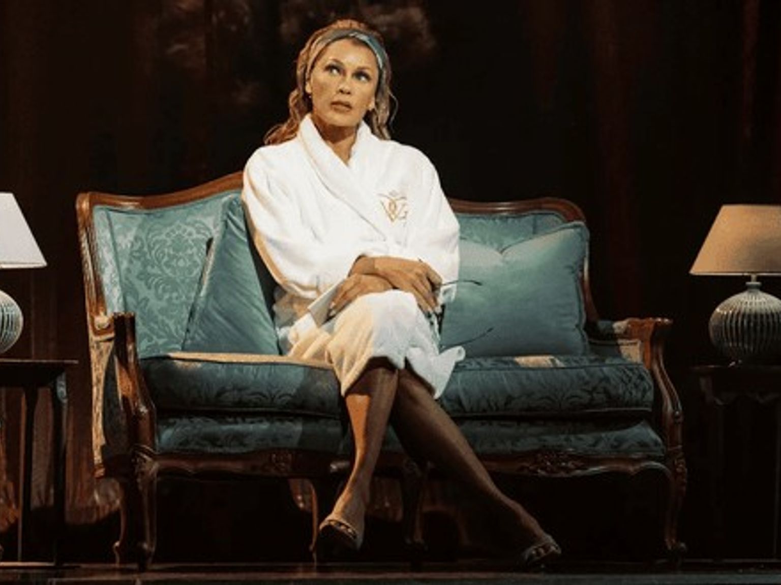 A woman in a white bathrobe and head wrap sits on an ornate teal couch on a dark stage set, looking up thoughtfully.