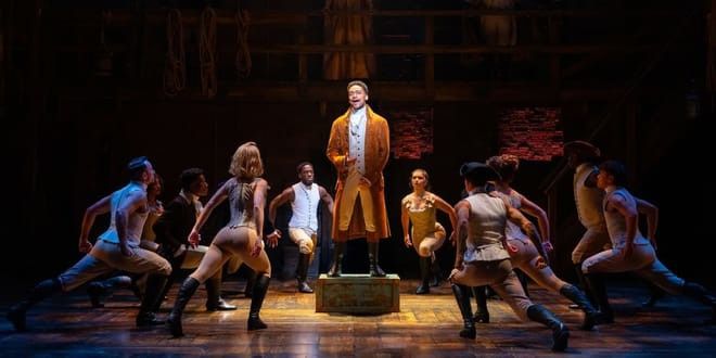 En artist i en orange rock står på en låda och sjunger på scen, omgiven av ett ensemble i tidstypiska kläder som intar dynamiska poser. Musikal – Hamilton – London.