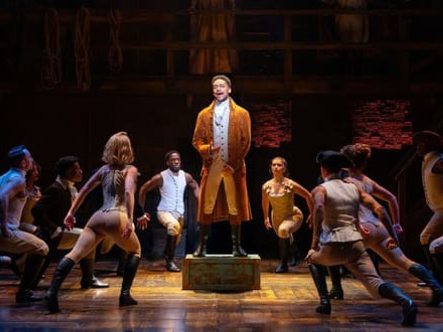 En artist i en orange rock står på en låda och sjunger på scen, omgiven av ett ensemble i tidstypiska kläder som intar dynamiska poser. Musikal – Hamilton – London.
