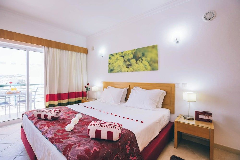 Ett hotellrum med en stor säng dekorerad med röda rosenblad och handdukar med texten "Muthu Hotel", vänd mot en balkong med utemöbler.