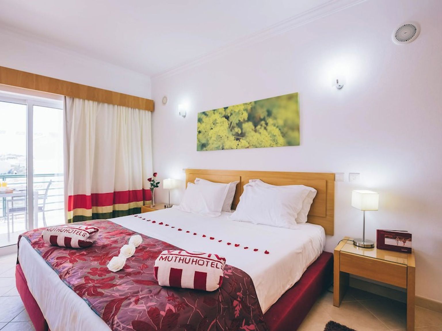 Ett hotellrum med en stor säng dekorerad med röda rosenblad och handdukar med texten "Muthu Hotel", vänd mot en balkong med utemöbler.