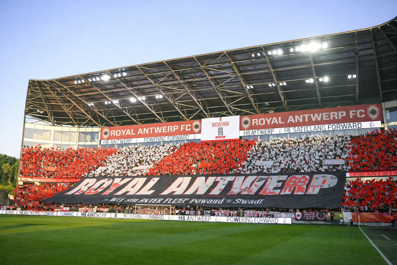 Fans op de tribunes van een voetbalstadion houden rode en witte kaarten omhoog en vormen een mozaïek achter een groot spandoek met de tekst "ROYAL ANTWERP".