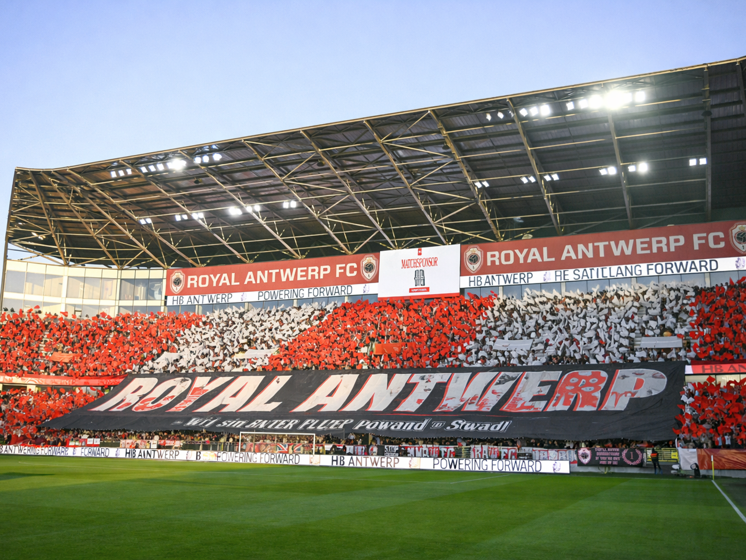 Fans in einem Fußballstadion halten rote und weiße Karten hoch und bilden ein Mosaik hinter einem großen Banner mit der Aufschrift „ROYAL ANTWERP“.