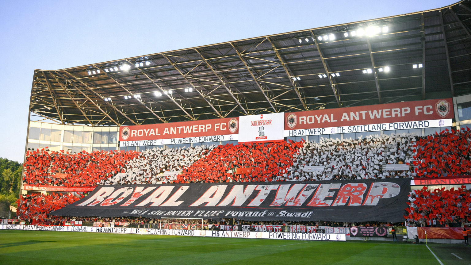 Fans op de tribunes van een voetbalstadion houden rode en witte kaarten omhoog en vormen een mozaïek achter een groot spandoek met de tekst "ROYAL ANTWERP".