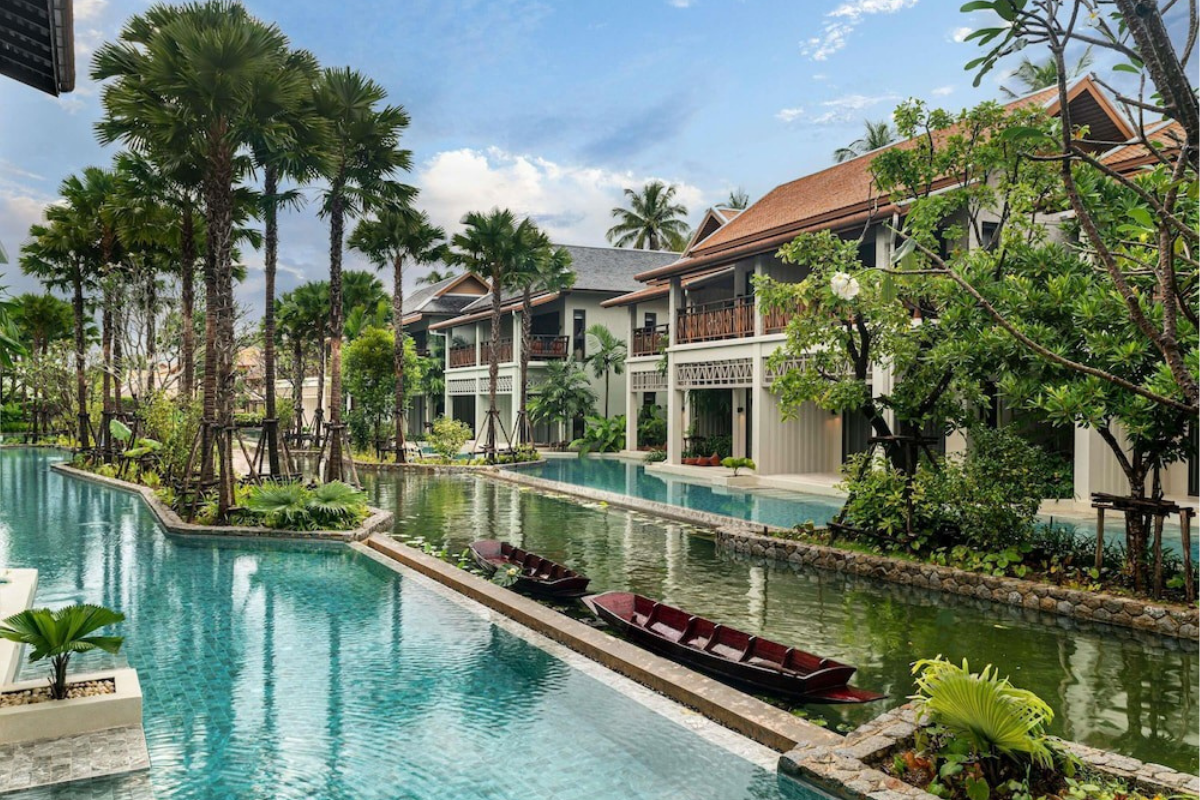 En tropisk resort med traditionella byggnader i thailändsk stil, slingrande pooler, palmer och två små träbåtar.