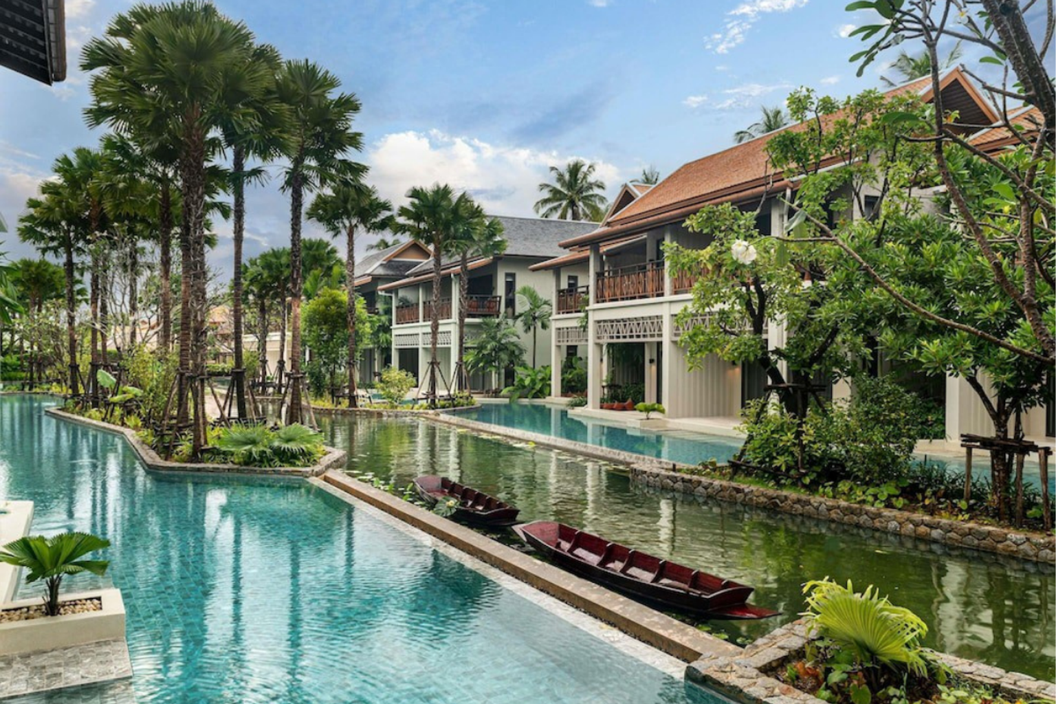 En tropisk resort med traditionella byggnader i thailändsk stil, slingrande pooler, palmer och två små träbåtar.