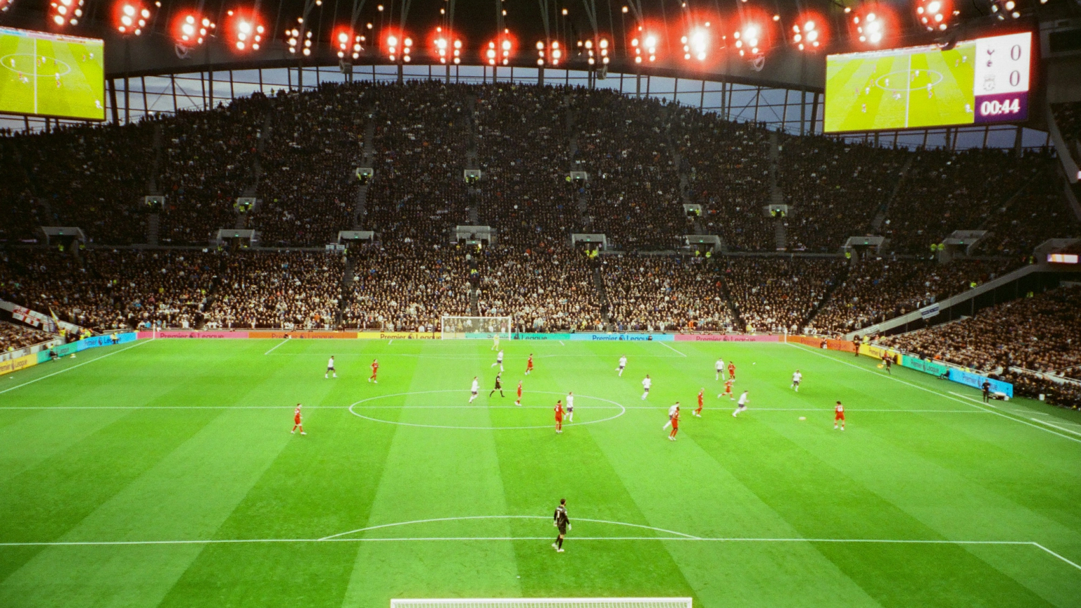 Widok z góry na mecz piłki nożnej na dużym stadionie z pełną widownią i tablicą wyników pokazującą 0-0.
