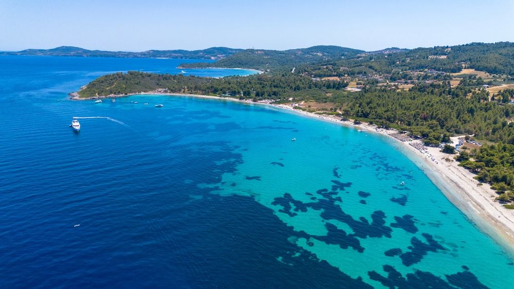 Luftfoto af et fantastisk kystlandskab med klart turkis vand, en sandstrand, grønne skove og en stor hvid yacht.