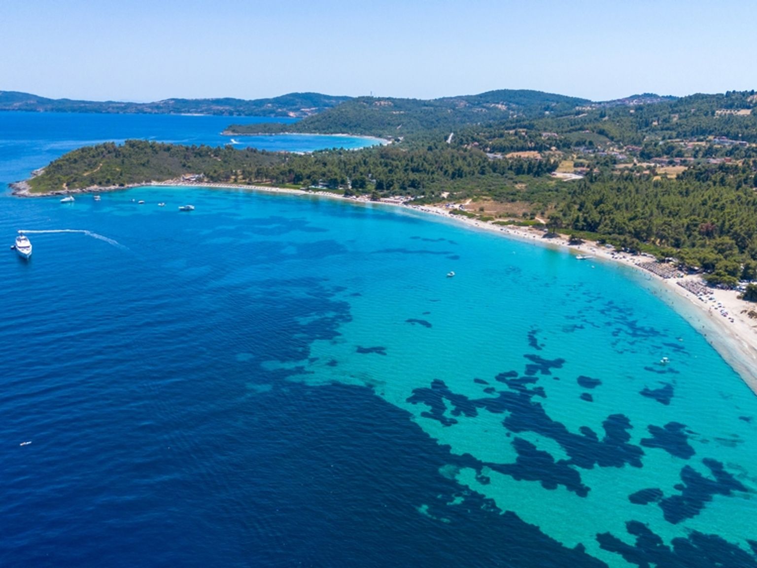 Luftfoto af et fantastisk kystlandskab med klart turkis vand, en sandstrand, grønne skove og en stor hvid yacht.